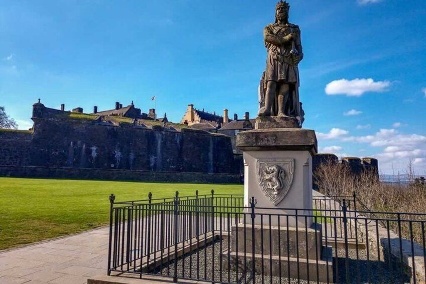 A statue in front of a historic stone wall with a grassy area and a clear blue sky. 2