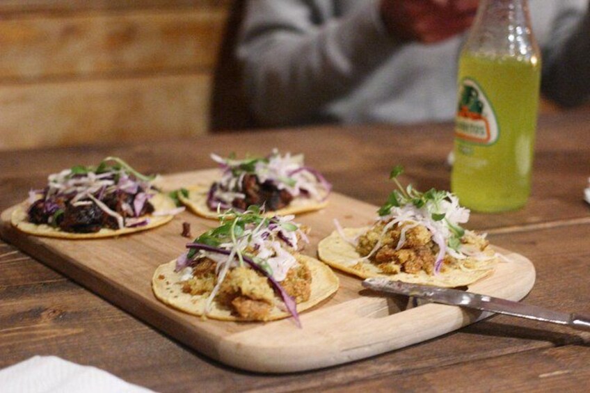 Et træbræt med tre tacos fyldt med kød og lilla kål, en flaske grøn drik og en kniv på et træbord. 2