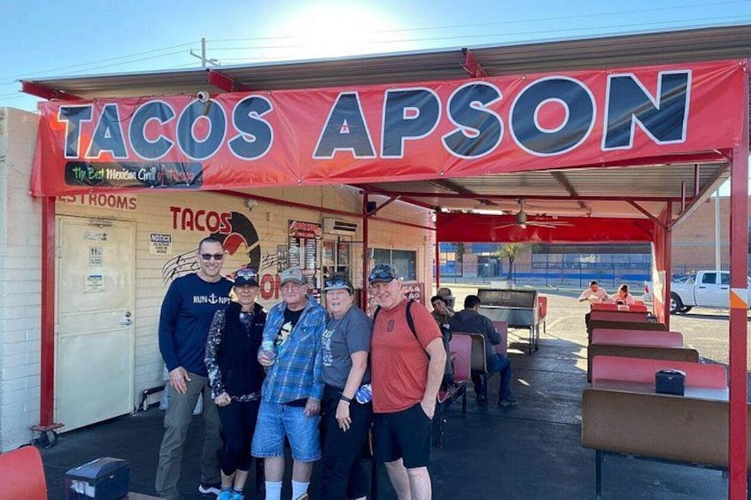 Un camión de comida llamado 'Tacos Apson' con un toldo rojo, un grupo de personas de pie al frente y un área de estacionamiento al fondo. 2