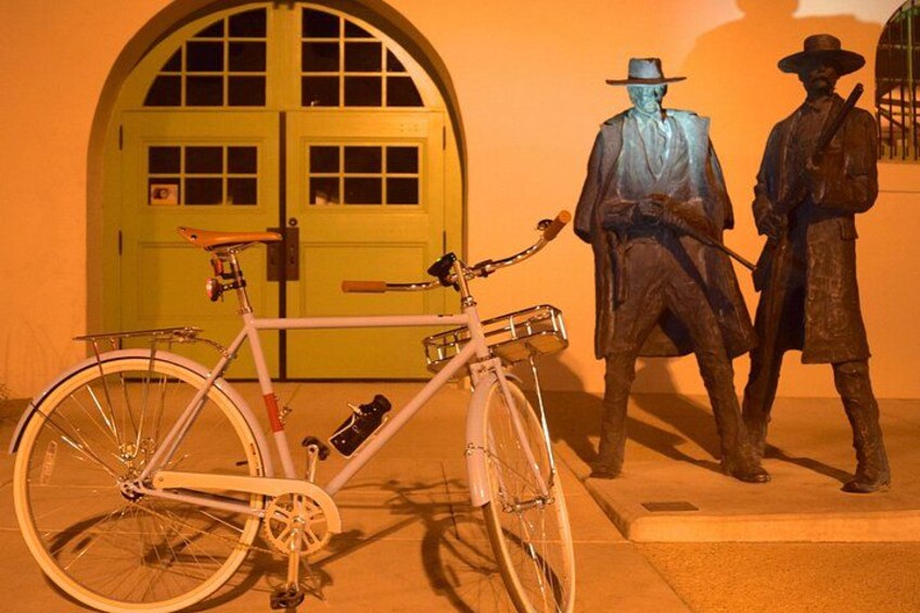 Una bicicleta blanca con canasta y botella de agua, una puerta verde y dos estatuas de bronce. 3