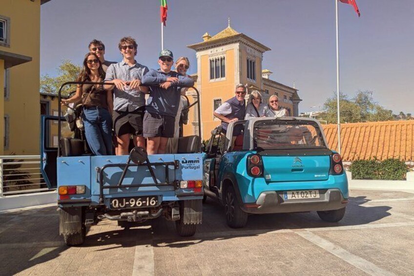 En grupp människor står framför en blå Portugal Turismo-bil med en liten släpvagn, parkerad framför en gul byggnad med en flagga. 5