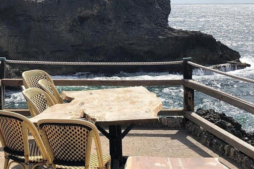 A wooden table and wicker chairs on a rocky seaside platform with a railing. 4