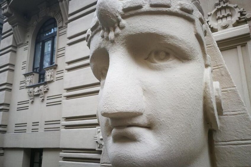 En stenskulptur af et ansigt med en detaljeret hovedbeklædning, foran en bygning med klassiske arkitektoniske træk. 1