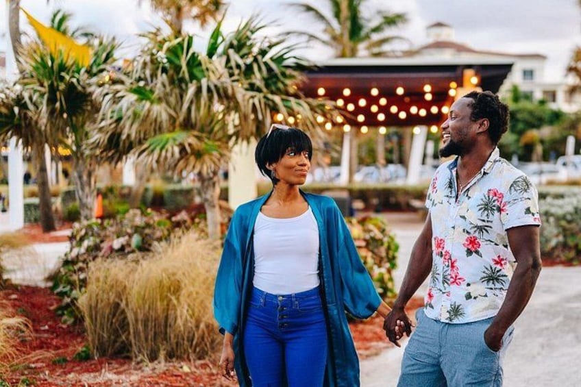Two people walking outdoors, palm trees, and a building with string lights in the background. 5