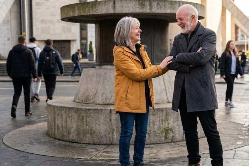To personer står i en offentlig plads med et rundt monument i baggrunden. 2