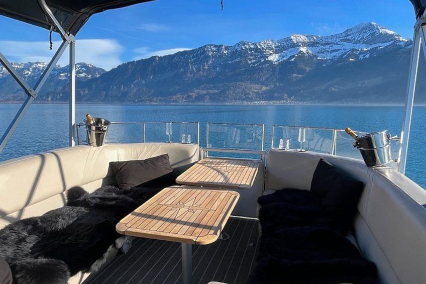 Cubierta de barco con una mesa y sillas, un asiento cubierto de piel, y vistas de montañas nevadas al otro lado del agua. 4