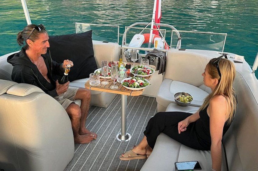 Un barco con dos personas sentadas en la cubierta, una mesa con comida y bebidas, y una masa de agua al fondo. 5