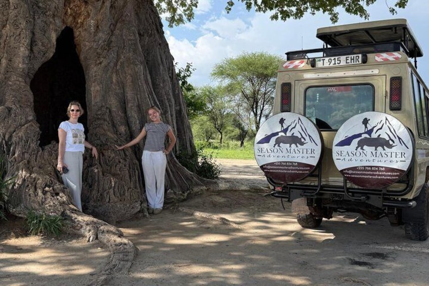 Two people standing next to a large tree with a vehicle labeled 'T-915 EHR' and 'Season Master Adventures' decals. 3