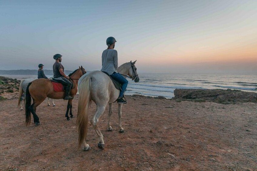 Three people riding horses on a dirt path by the sea during sunset. 1