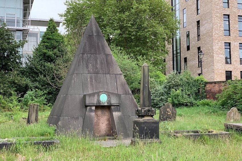 Een piramidevormig monument, omringd door begroeiing en grafstenen. 1
