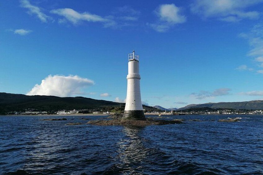 A white lighthouse on a rocky outcrop surrounded by water, with a clear sky and distant hills. 1