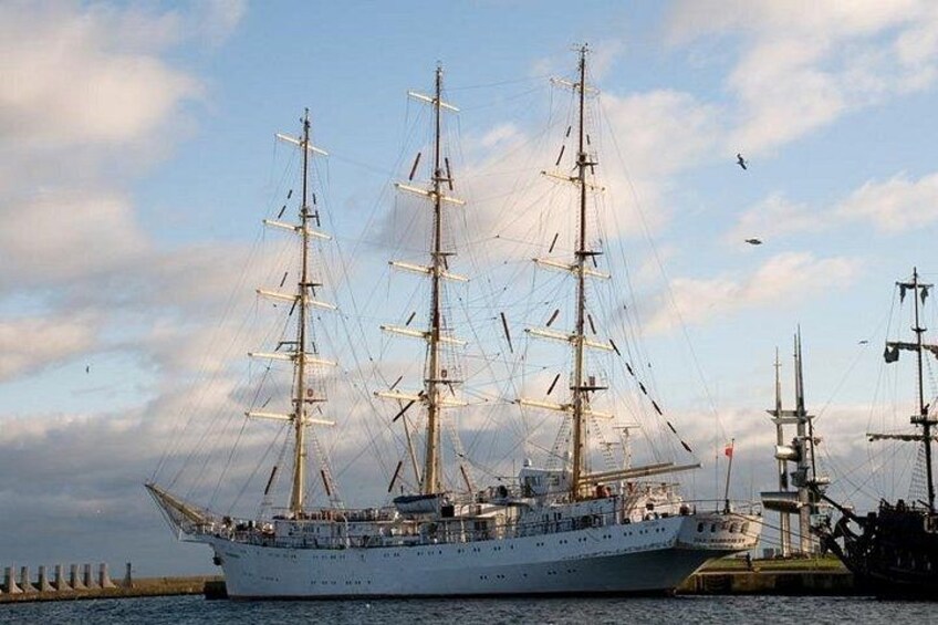 Un gran barco de vela con varios mástiles y velas, atracado en un puerto con otros barcos y un rompeolas al fondo. 5