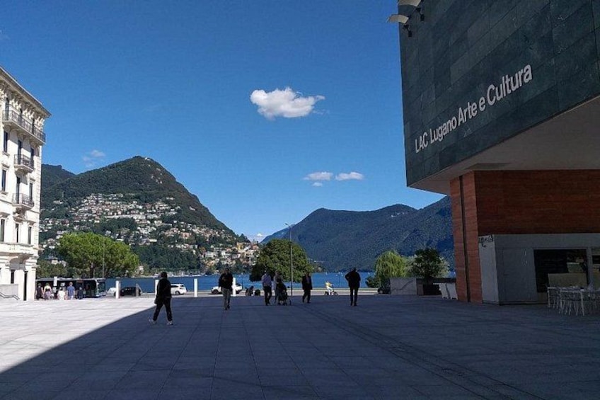 A plaza with people walking, a mountain in the background, and a building with 'LAC Lugano Arte e Cultura' sign. 1