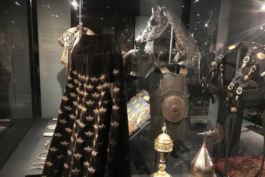 En udstilling af historiske genstande, herunder en sort kjole med gyldne kroner, en hjelm og en heste statue. 3