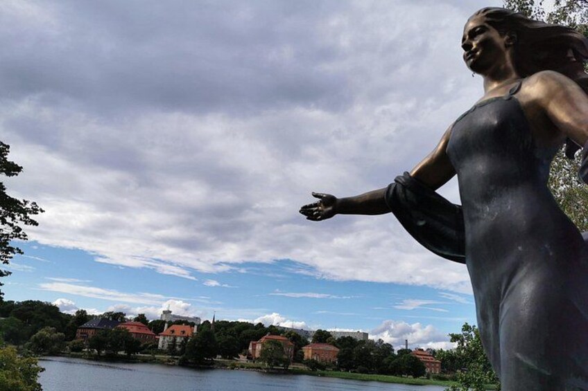 En statue af en kvinde i kjole ved en sø, med bygninger i baggrunden. 2