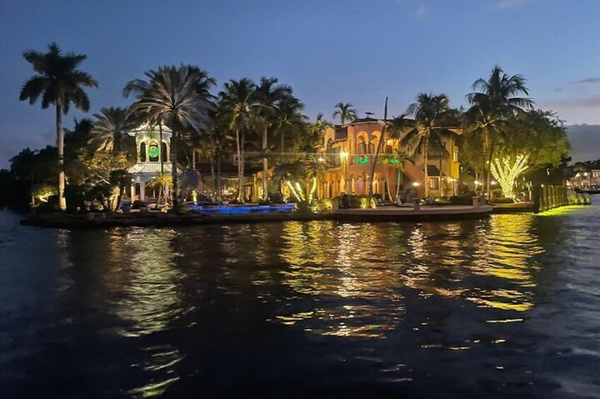 A waterfront with illuminated buildings, palm trees, and a calm body of water reflecting the lights. 1