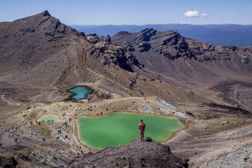 A person standing on a rocky outcrop overlooking green lakes and a mountainous landscape. 1