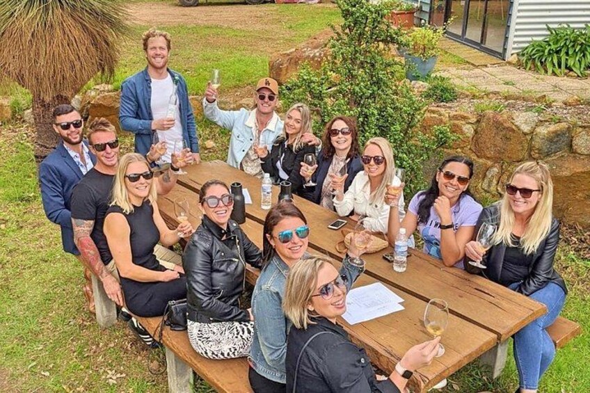 A group of people sitting around a wooden table outdoors, holding drinks. 2