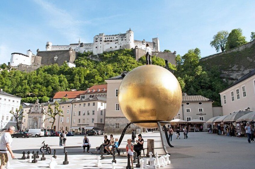 A large golden sphere in an open square with historic buildings and a castle in the background. 3