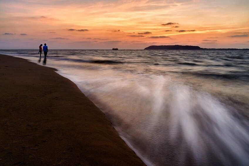 A beach with two people walking along the shore, a distant island, and a boat on the water. 3