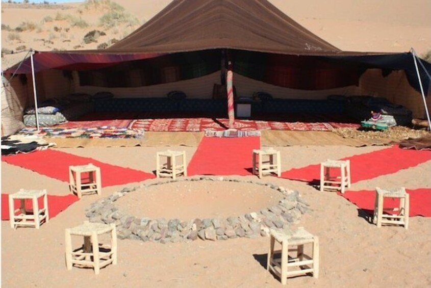 A desert campsite with a large tent, red carpets, and wooden stools. 4