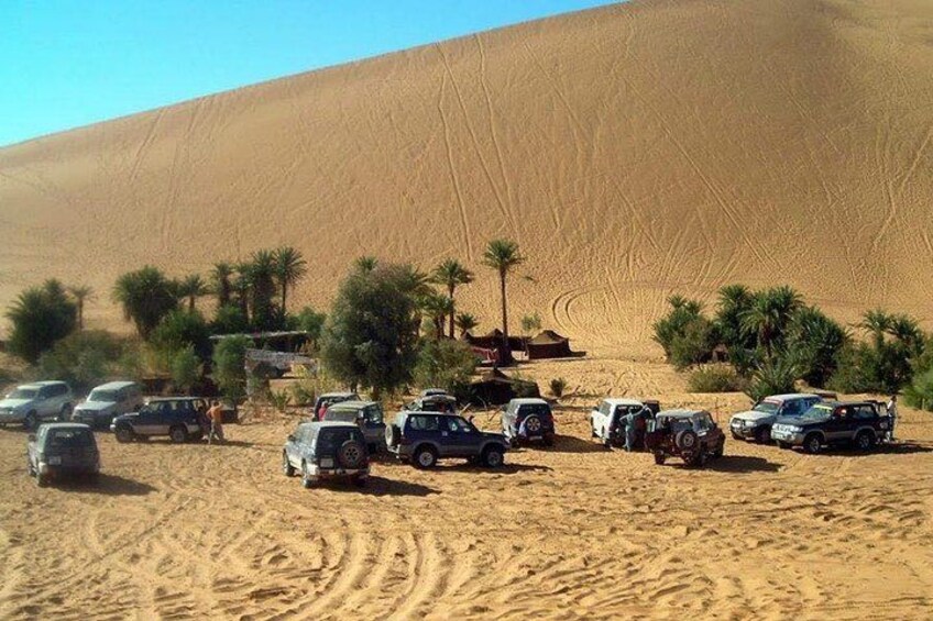 A desert landscape with sand dunes, palm trees, and a group of vehicles parked on the sand. 5