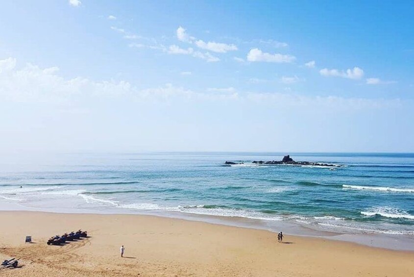A sandy beach with waves, a person walking, and a rock formation in the distance. 2