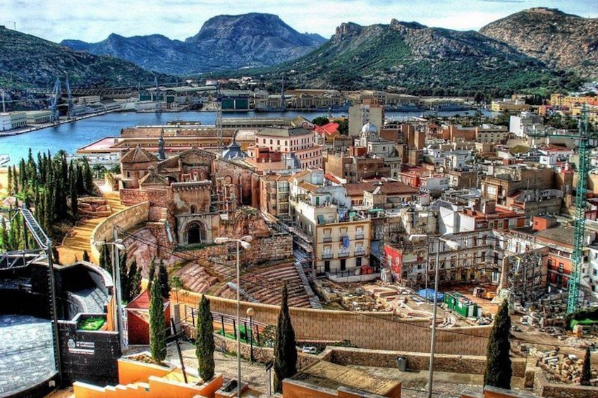 Un paysage urbain côtier avec un amphithéâtre historique, un port et, au loin, un paysage montagneux. 2