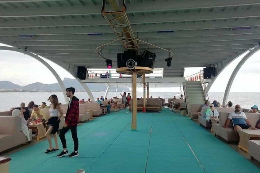 Ein Bootsdeck mit sitzenden und stehenden Personen, einer Bühne mit Lautsprechern und Blick auf das Meer. 2