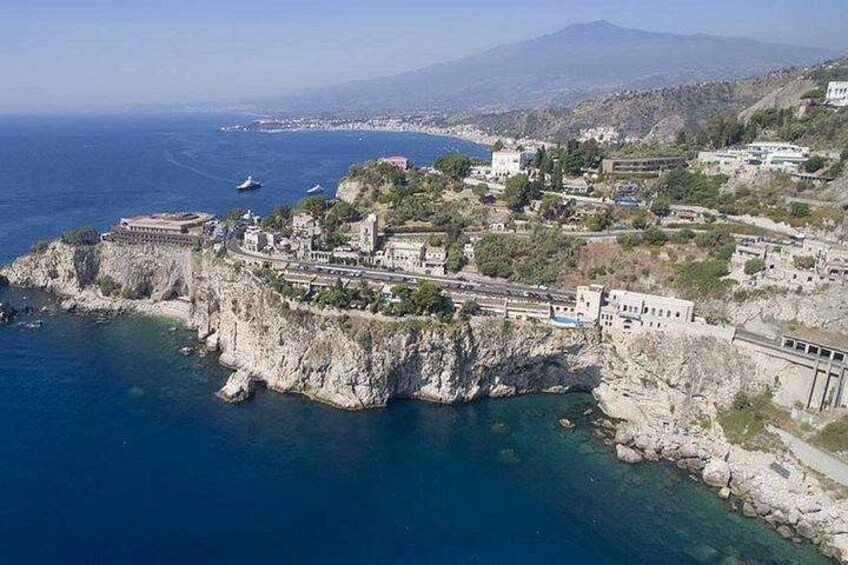 A coastal town with buildings on a cliff, a clear blue sea, and distant mountains. 1