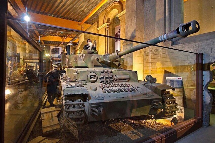 A museum exhibit of a military tank with a large gun, surrounded by glass display cases and a brick floor. 1