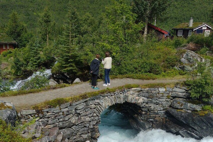 Een stenen brug over een waterval, omringd door weelderige begroeiing en een klein huisje. 5