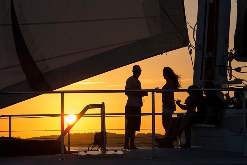 A sunset with a sailboat silhouette, people on the deck, and a railing. 2