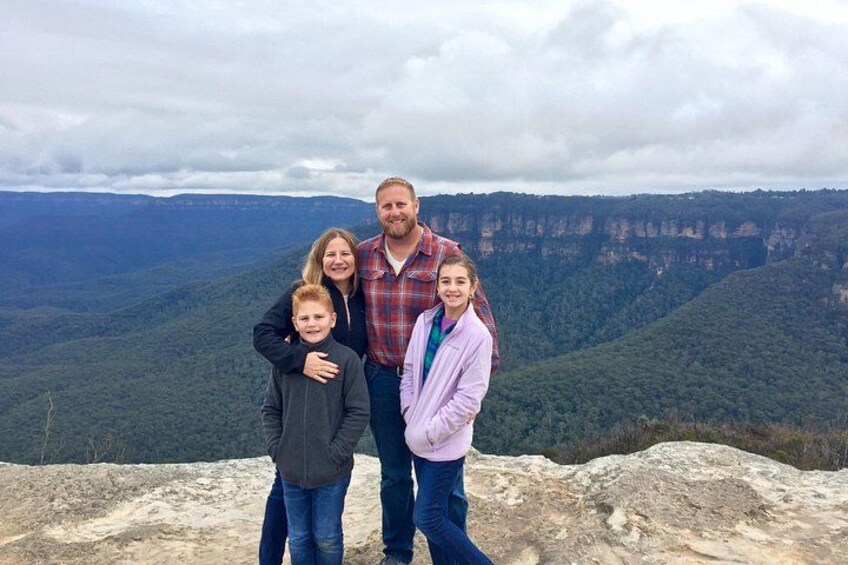 Una familia parada sobre un promontorio rocoso, con un paisaje montañoso al fondo. 3