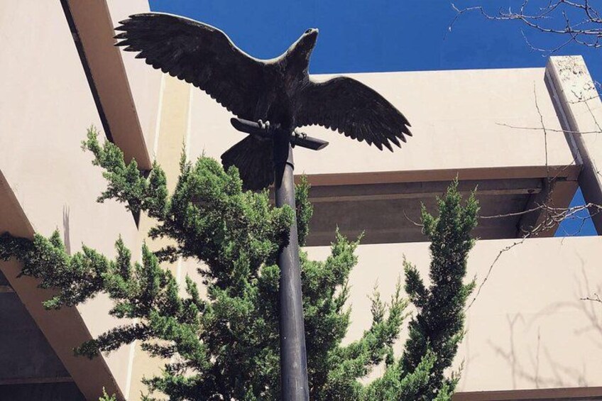 A statue of a bird with outstretched wings on a pole, surrounded by trees and a building. 3