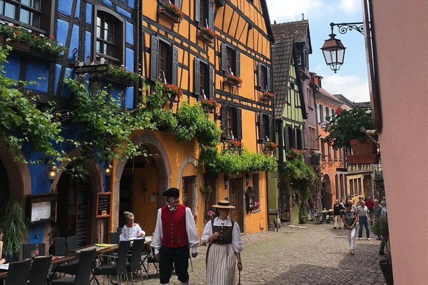 A cobblestone street lined with colorful half-timbered buildings, a person in traditional attire, and outdoor seating areas. 1