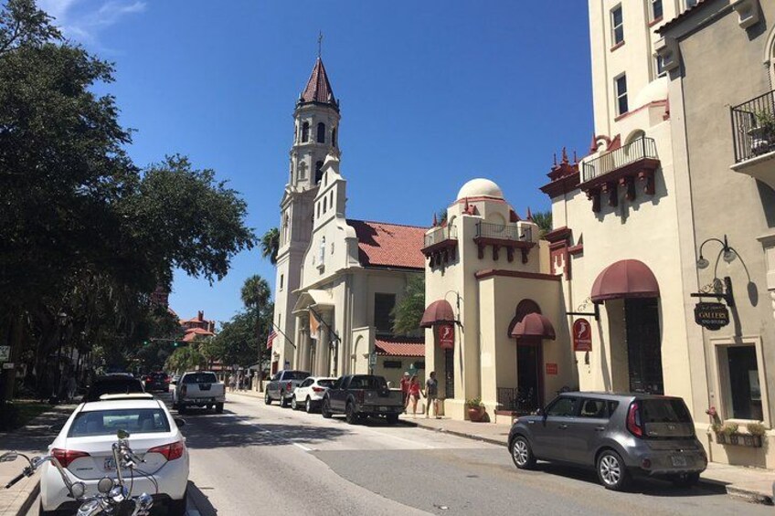 Una calle con coches aparcados, una iglesia con campanario y edificios con toldos rojos. 1