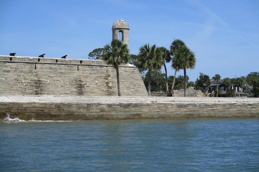 Un muro de piedra con una torre, palmeras y una masa de agua tranquila. 4