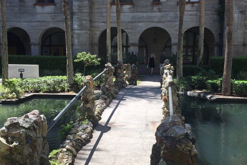Un puente de piedra sobre el agua, con un camino, rodeado de vegetación y un edificio con arcos al fondo. 3