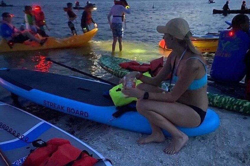 A person sitting on a blue inflatable board on a sandy beach, with kayaks and people in the background. 3