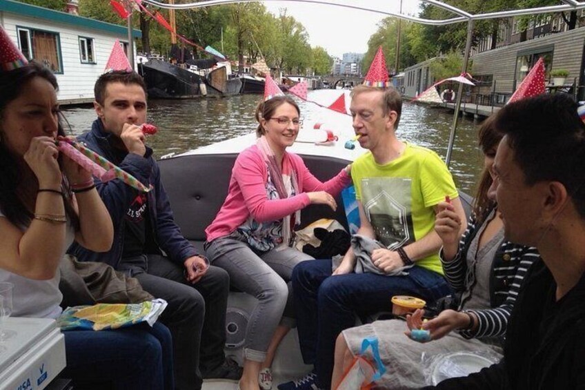 A group of people on a boat, some holding drinks and wearing party hats, with a river and houses in the background. 2