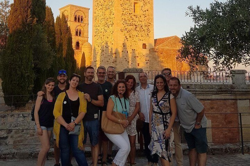 A group of people posing in front of a historic stone tower with a bell tower, surrounded by trees and a stone wall. 3