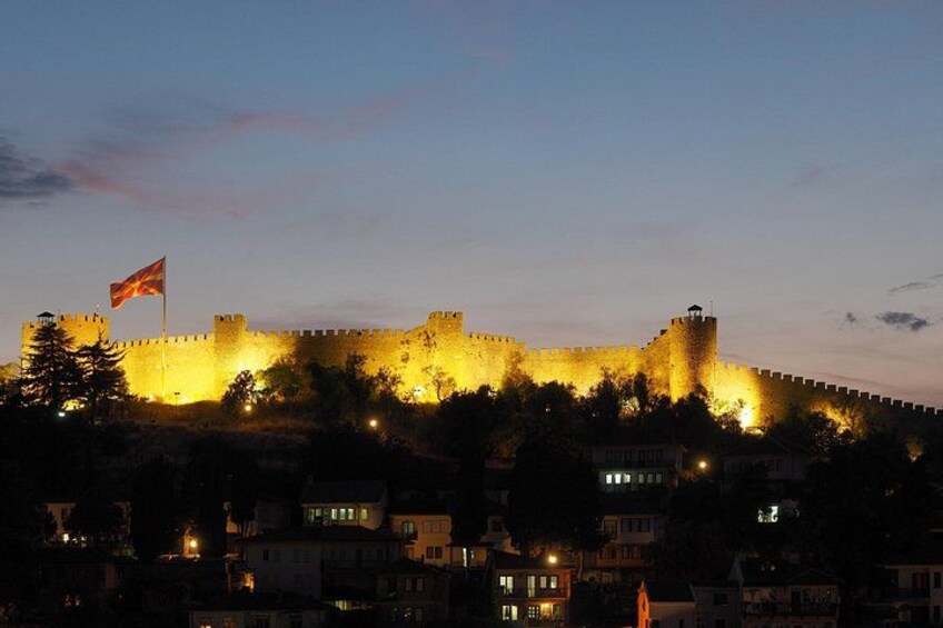 A fortified castle at dusk with a flag, surrounded by buildings and trees. 3