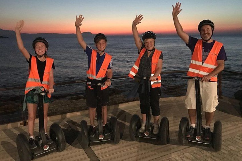 Vier mensen op segways met opgetrokken armen, zichtbaar aan hun veiligheidshesjes, aan een waterkant bij zonsondergang. 3