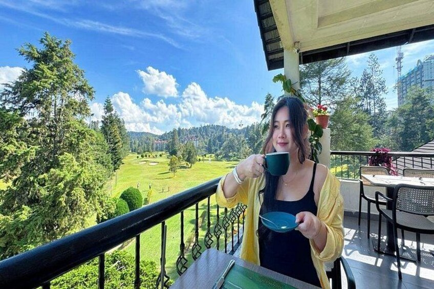 Una persona sosteniendo una taza, sentada en un balcón con vista a un paisaje verde y árboles. 3
