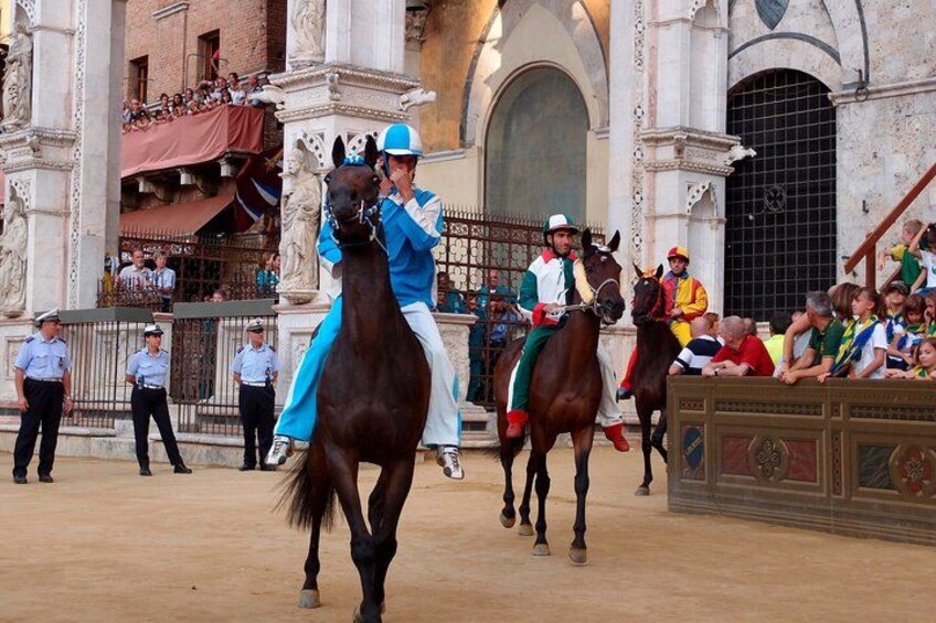 A horse riding event with participants in colorful attire, a crowd watching, and a historic building in the background. 1