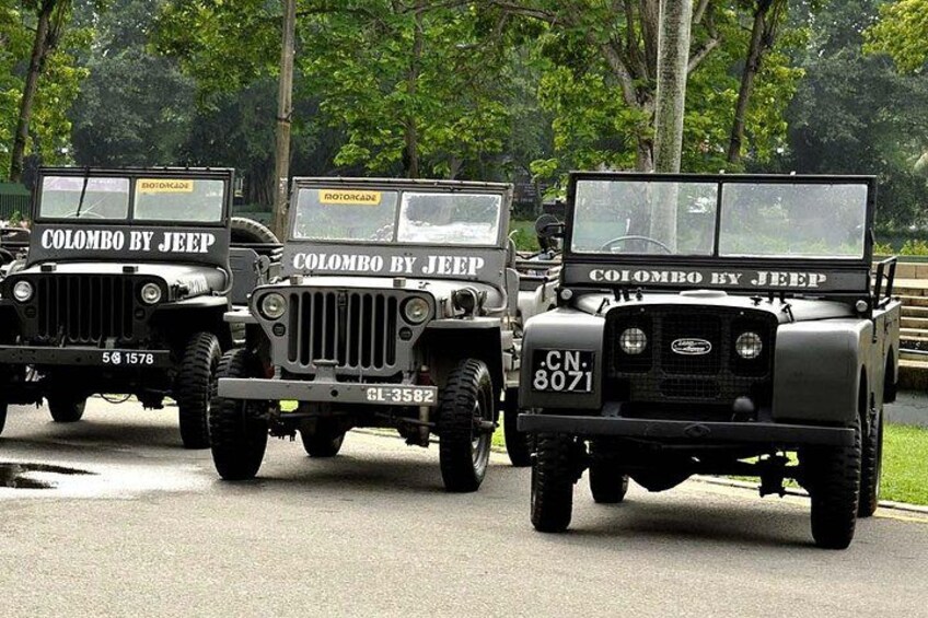 Three black military jeeps with 'Colombo by Jeep' written on them, parked on a road with trees in the background. 1
