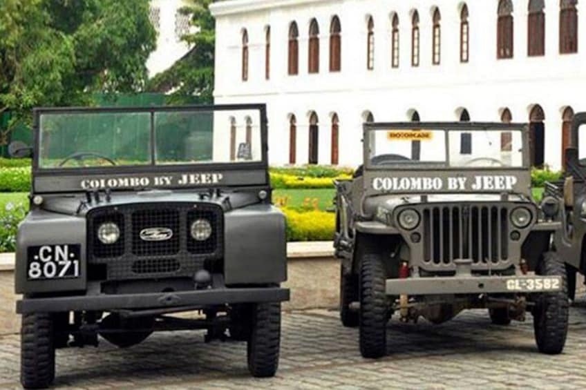 Dos jeeps militares con la frase 'Colombo by Jeep' escrita en ellos, estacionados frente a un edificio con ventanas de arco. 3
