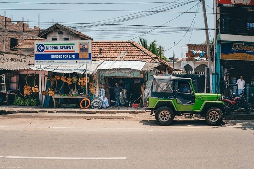 A green vehicle parked on a street, a shop with a sign for Nippon Cement, and a building with a sign for S-Ron. 2