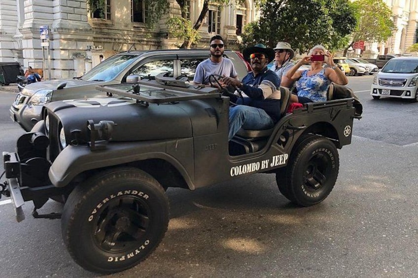 A military-style jeep with 'Colombo by Jeep' written on the side, parked on a street with buildings in the background. 4